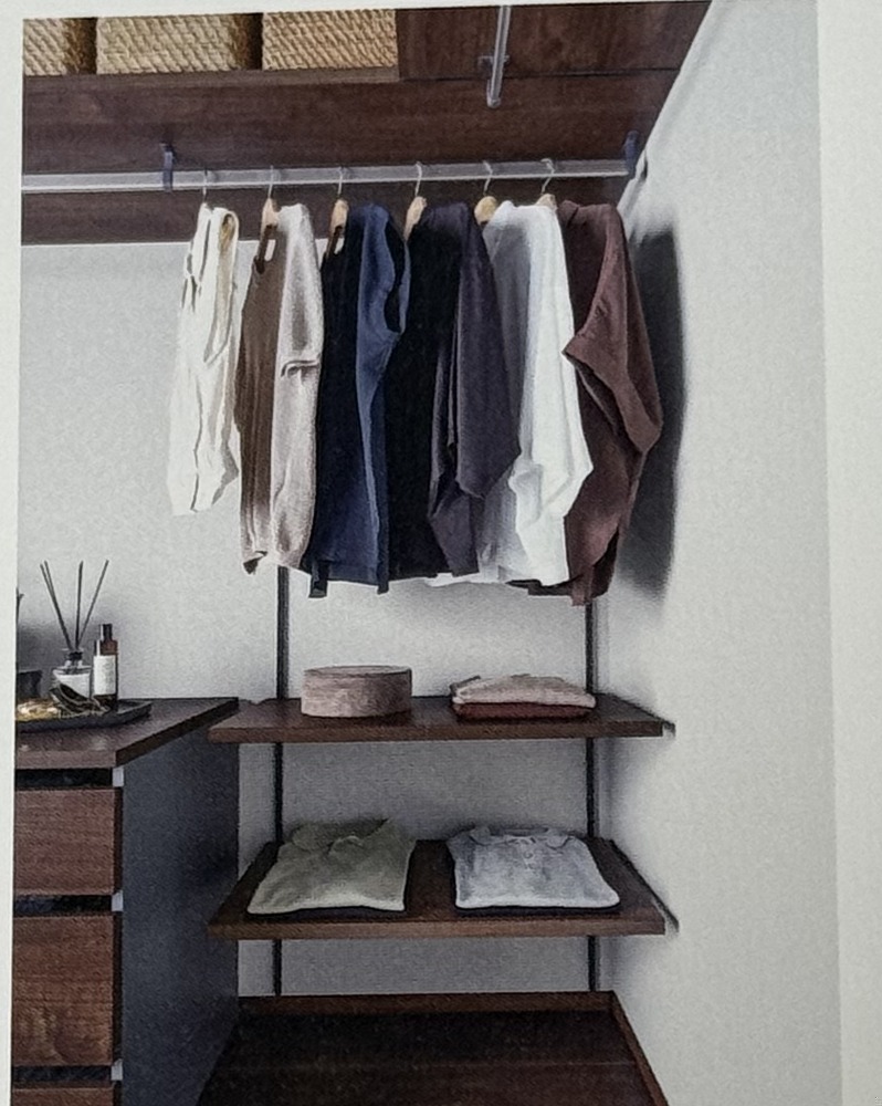 Next Walk-in Closetの背面自在棚。棚柱に取り付けられた濃い木目の棚板の上に小物やクッションが並び、下にハンガーで服が掛かっている